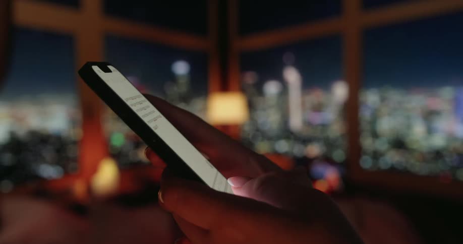 Closeup of hands texting on a smartphone in a dimly lit room with raincovered windows and city lights glowing in the background