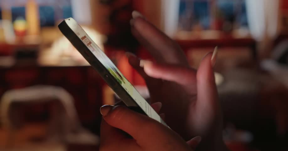A womans hand with manicured nails holding a slim smartphone at a sharp angle against a blurred warmtoned background
