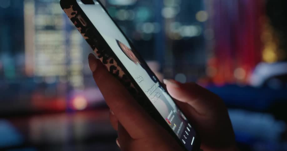 A person scrolls through social media on a smartphone with a leopard print case set against a vibrant cityscape glowing with colorful lights