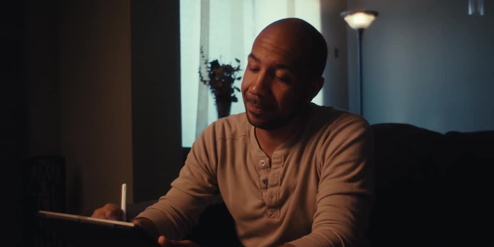 A man sitting on a sofa and looking at a tablet and moving his head from side to side in tones of disappointment