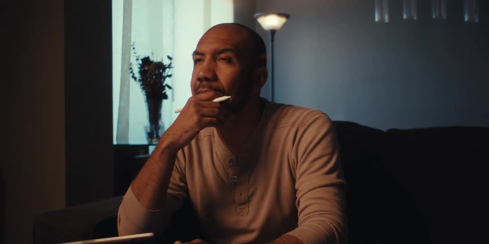 The face of a seated man who is with his hand on his chin in thoughtful tones and holding a white pen