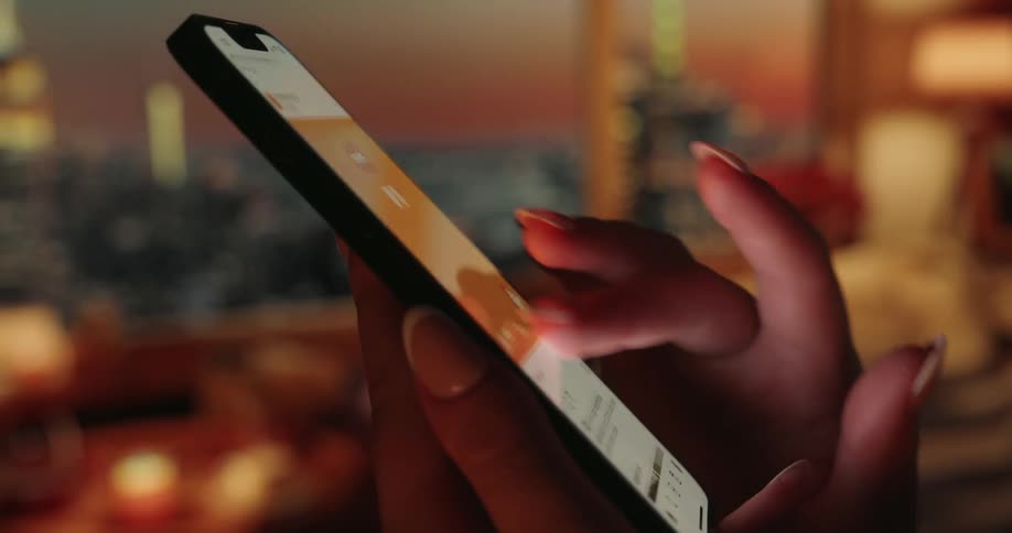 A hand scrolls on a phone screen in a closeup with a window revealing a cityscape at sunset in the background