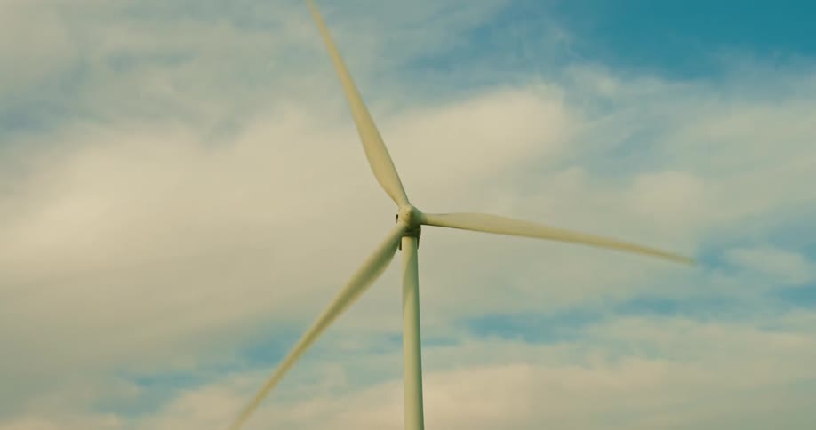 A closeup captures the rapid blurred motion of a wind turbines massive white blades as they spin against a backdrop that has white fluffy clouds going across a light blue sky