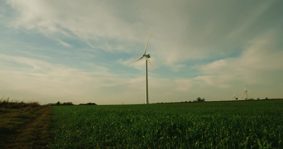 From a vantage along a path cutting through farmland a tall wind turbine stands in a lush green field its blades slowly rotating beneath a partly cloudy sky while the surrounding open landscape extends beyond the horizon
