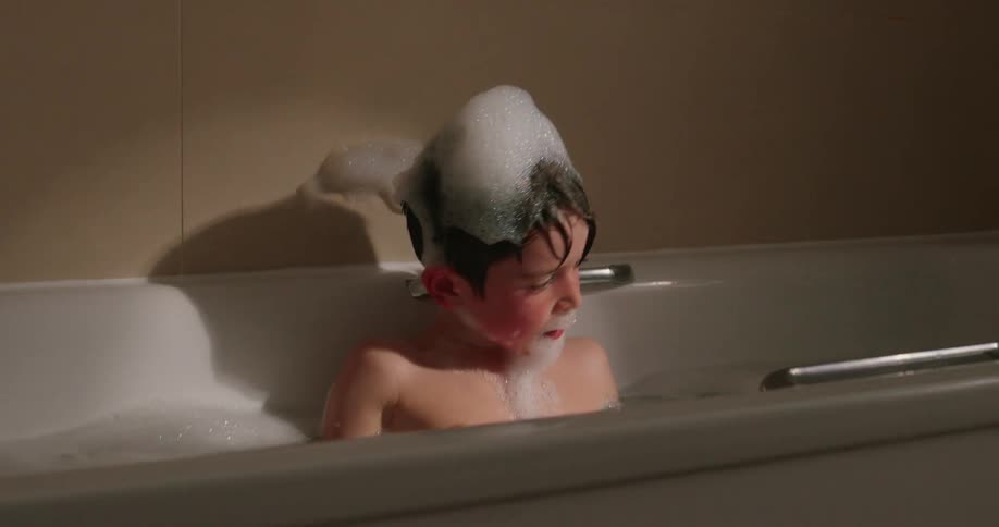 Young boy with sud on his head taking a bubble bath in a white bathtub