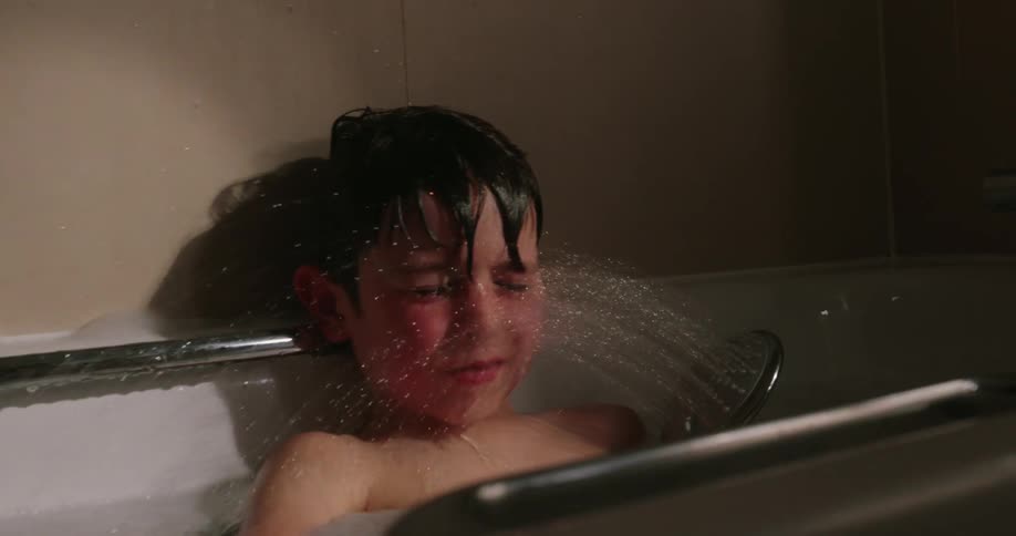 A boy takes a bath in a tub using a handheld showerhead to spray water