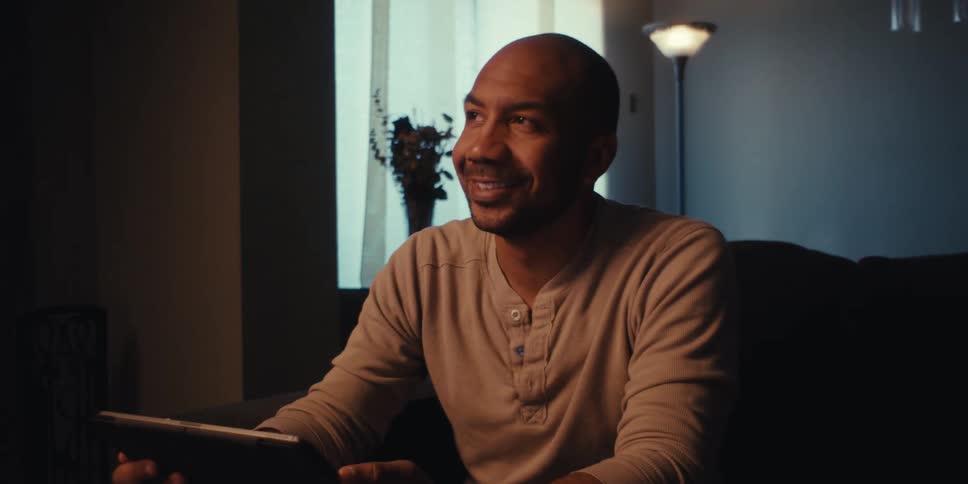 A middleaged man with a shaved head and a short beard sitting in a dimly lit room wearing a beige henley shirt holding a tablet and a stylus while looking up with a thoughtful expression with a vase of flowers