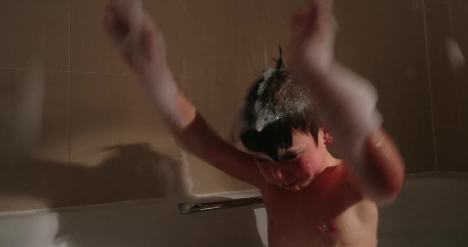 A boy plays with foam in a bathtub throwing it into the air with his arms in a bathroom