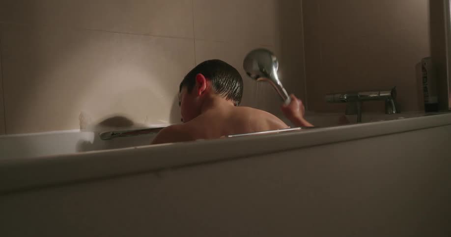 A kid taking a bath in a bathroom holding a hand shower spraying water with his back turned to the camera