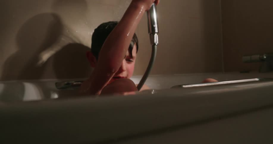 A boy in a bathtub sprays water over his head using a handheld showerhead in a bathroom
