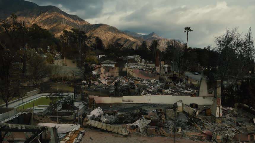 The charred remains of a destroyed house stand amidst a landscape of burnt trees and rubble with mountains looming in the background under a cloudy sky