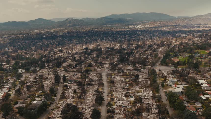 An aerial view shows a neighborhood devastated by fire with charred remains of houses and trees amidst a vast landscape leading to distant mountains