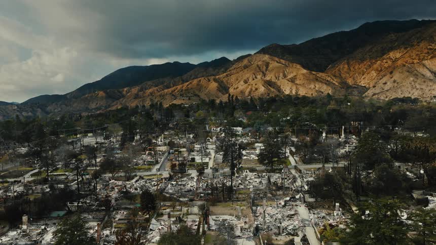 A neighborhood lies in ruins reduced to rubble and charred remains against a backdrop of mountains under a stormy sky