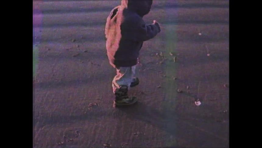 A child in a hoodie gathers pebbles from the wet sand on a beach illuminated by the warm hues of a sunset