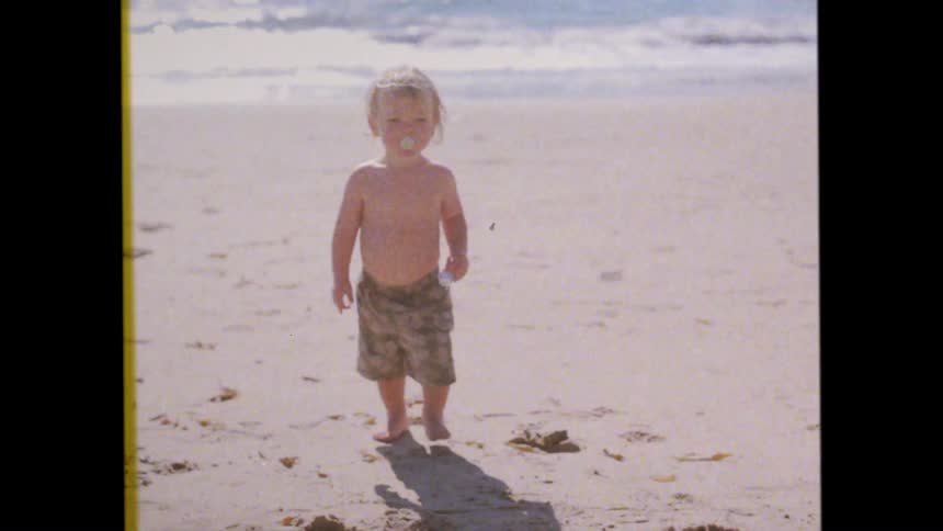 A toddler with light hair and wearing patterned shorts stands barefoot on a sandy beach with the ocean and waves visible in the background