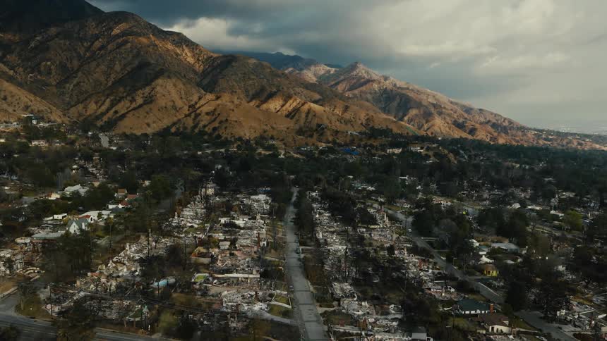 An aerial drone surveys a residential landscape ravaged by wildfire displaying complete destruction of homes and scorched foliage under a somber sky as it sweeps over the area mountains can be seen in the back
