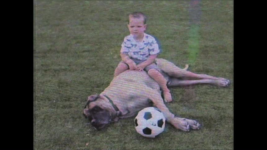 The little boy is sitting on top of the dog that is laying in the grass on its side