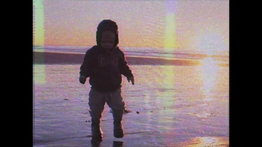 A child wearing a hoodie plays on the wet sand of a beach at sunset