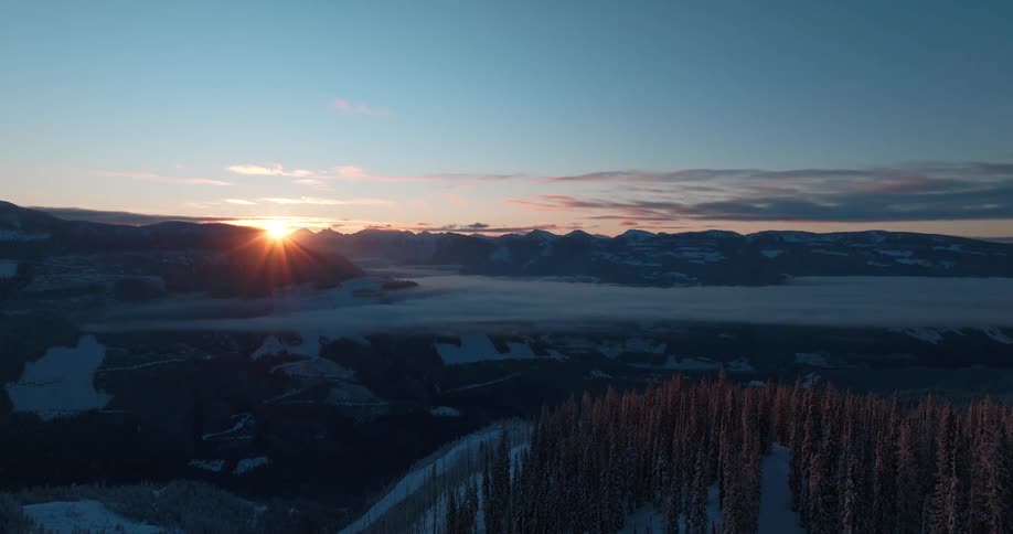 A sunrise peeks over distant mountain peaks casting a warm glow on a snowy landscape below