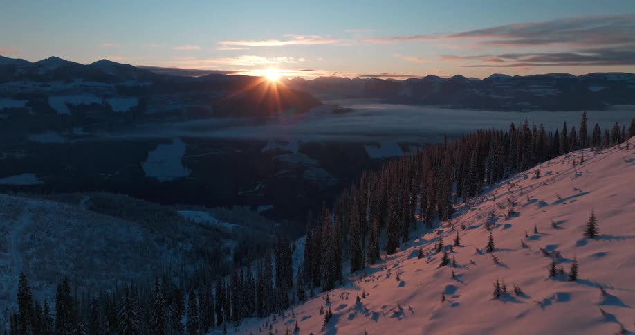 The sun is rising over snow and forest covered mountains