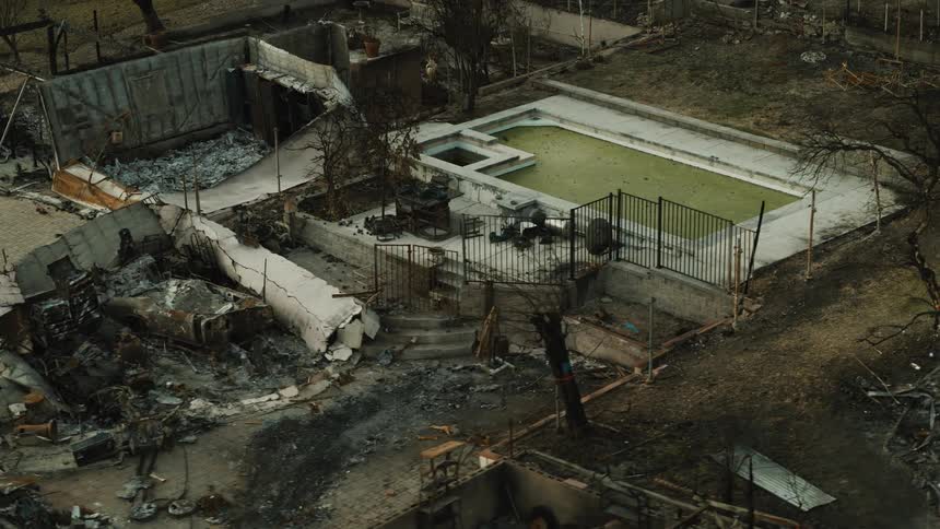 An aerial view shows a destroyed building next to a stillintact swimming pool with green water amidst a landscape of charred debris