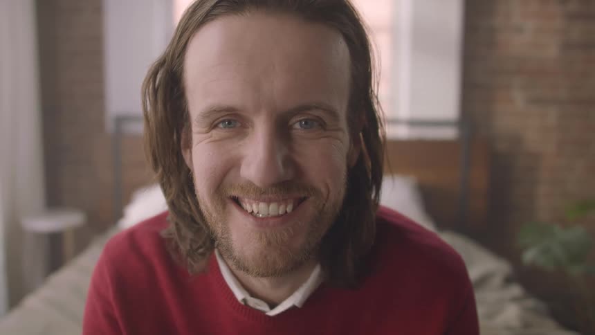 A closeup shows a smiling face with long blonde hair and a beard sitting on a bed at home looking at the camera