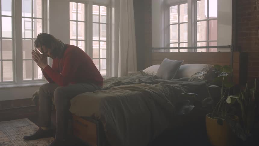 A person sits at the edge of a bed in a room adorned with indoor plants and illuminated by large windows