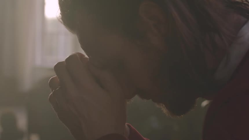 A closeup profile of a man with a beard and long hair his hands clasped against his forehead seated by a window overlooking a city in a dimly lit room