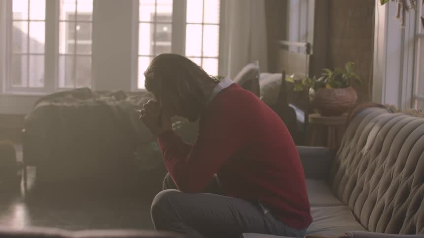 A person with long hair sits on a couch with their head down and hands clasped
