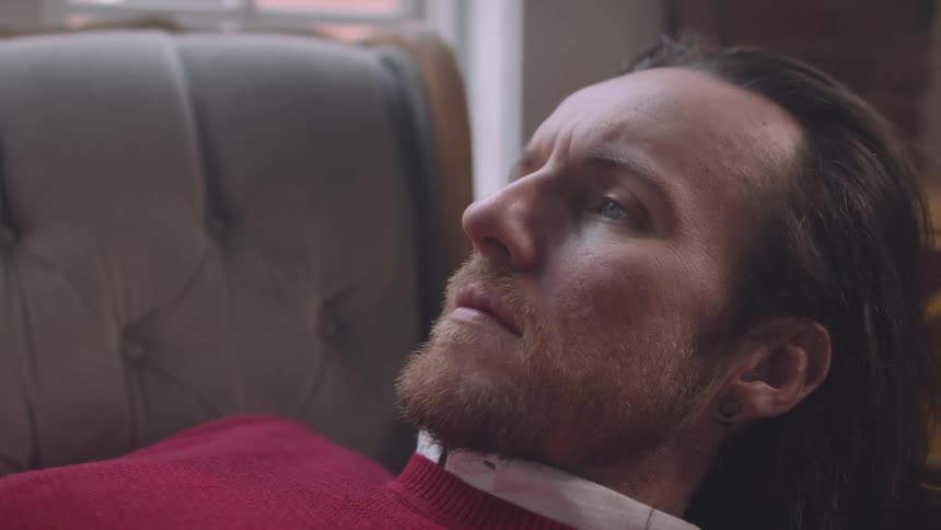 A closeup shows a man with long hair and a beard resting on a couch staring thoughtfully as if deep in contemplation