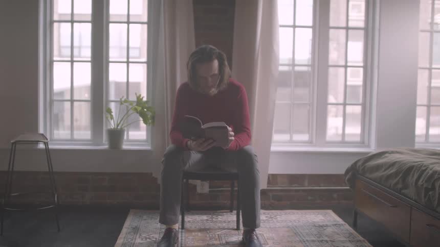 Man sitting in a char near a large picture window reading a book