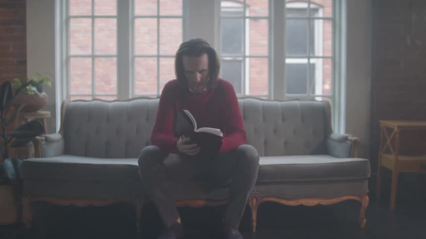 A person sits on a vintagestyle sofa in a room with large windows exposed brick walls and soft natural light engrossed in reading a book