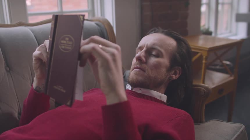 Man in red sweater laying on couch reading an old hardcover book
