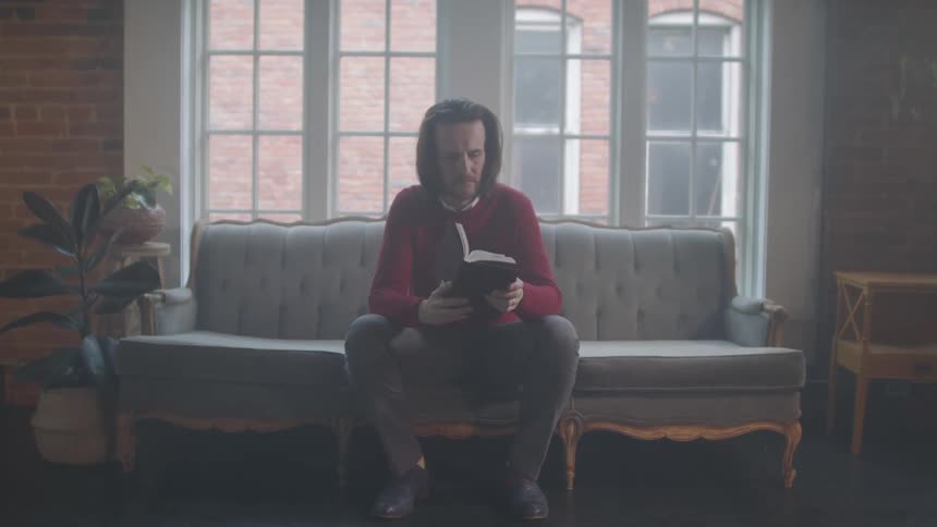 Man sitting on a couch near a large picture window reading a book