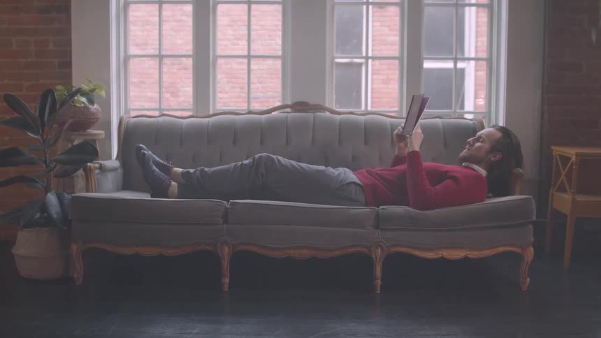 Man lies on a couch near the window while reading a book