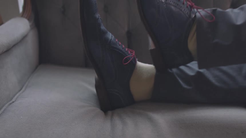 A pair of legs in gray pants and blue dress shoes with red laces rests on a gray upholstered couch