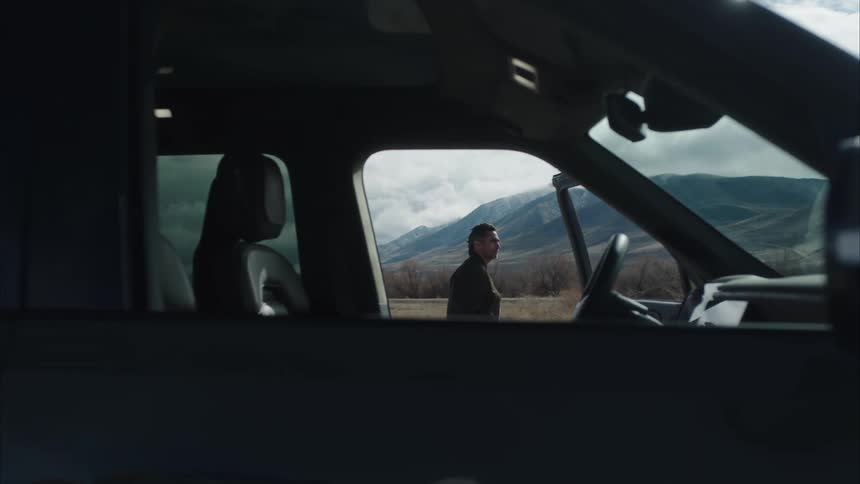Man stands in a field near a parked vehicle with woods and mountains in the background