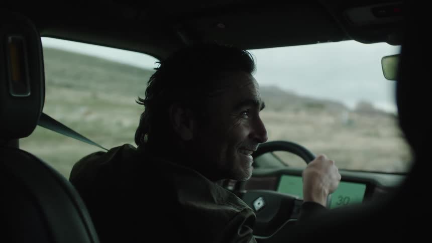A man with dark hair and a dark jacket smiles and looks to the side while driving a vehicle with a dashboard visible through a landscape with hills and a cloudy sky