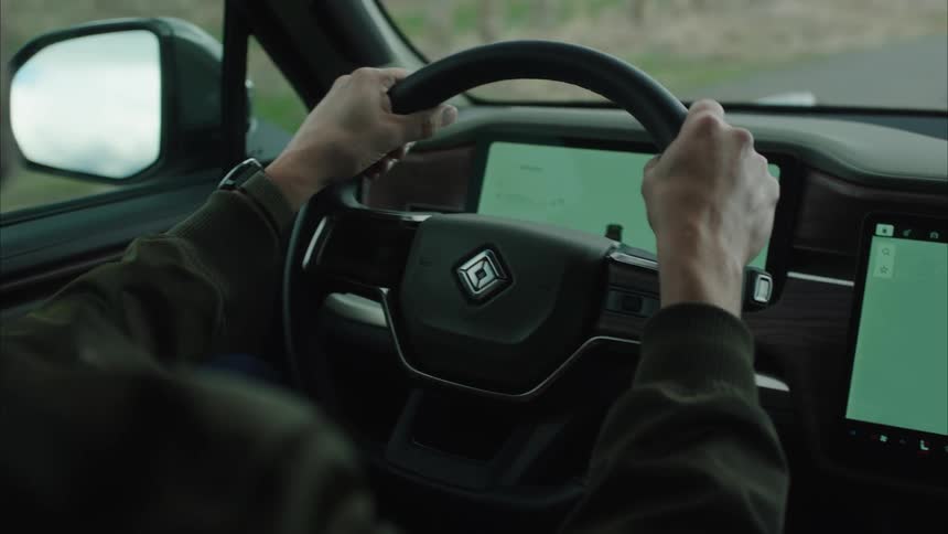 The hands of a person holding a black steering wheel in there is a logo in center of it