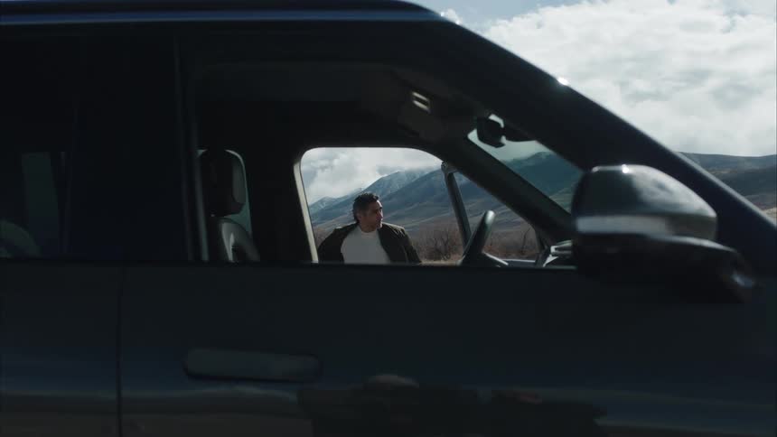 A person about to board is visible through the open drivers side window of a dark vehicle with a mountainous landscape in the background