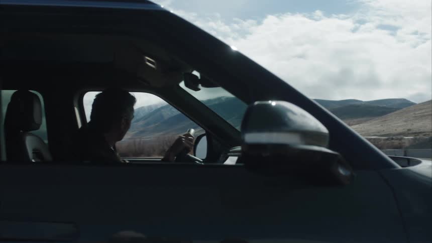 A man seated in the drivers seat of a car steps out to observe a landscape of barren mountains under white clouds