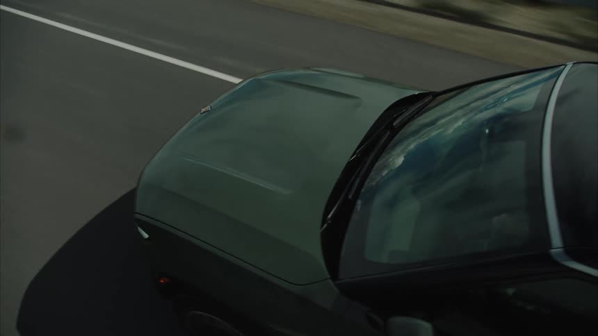 A dark suv drives down a straight asphalt road adjacent to a large body of water with distant mountains visible under an overcast sky