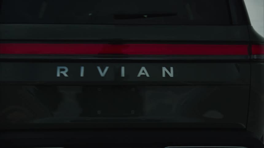 A dark rivian r1s travels along a paved road through a vast lakeside landscape beneath an overcast sky