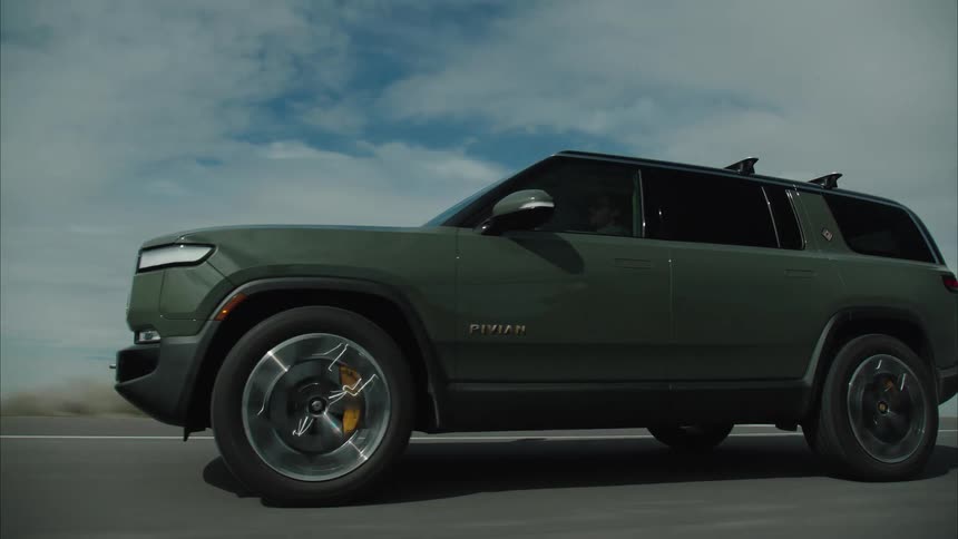 A dark rivian r1s travels along a paved road through a vast lakeside landscape beneath an overcast sky