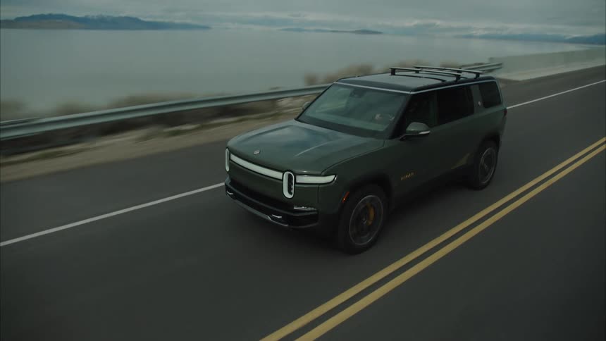 A dark green rivian suv with a roof rack drives along a highway next to a large body of water under an overcast sky