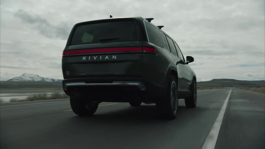 The suv is going down the road and in the back ground there are snow capped mountains with water at the base of them