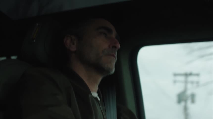 A man with a thoughtful expression is driving a car on a dull day gazing upwards with a slight smile