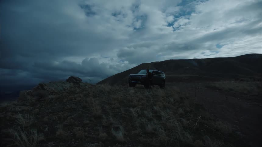 A man gets out of a black suv that is parked on the side of the road infront of a mountain and he walks to the edge of the cliff and stands there