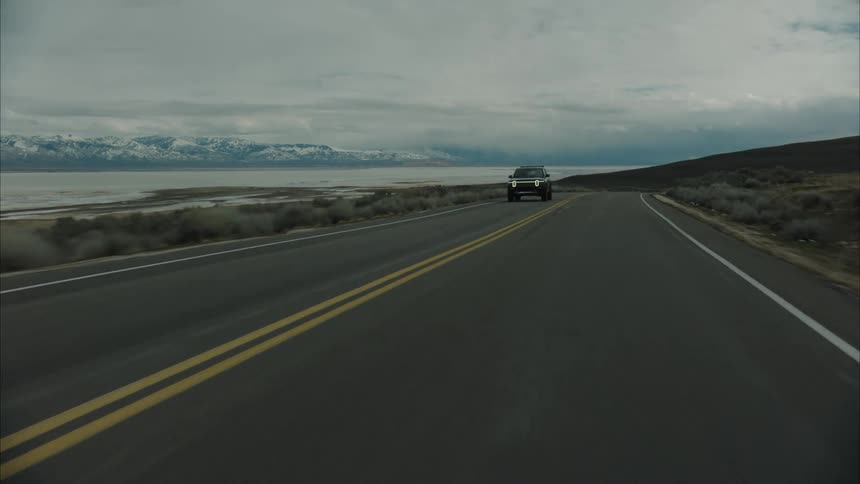 A darkcolored rivian r1t drives down a straight asphalt road with a lake and snowcapped mountains in the distance under a cloudy sky