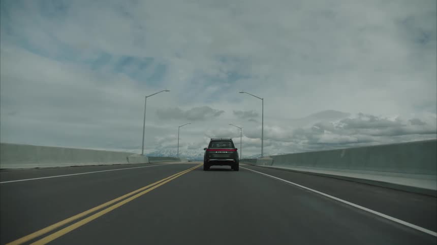 A dark rivian suv drives along a straight road with yellow lines towards a distant body of water and mountains under a cloudy sky
